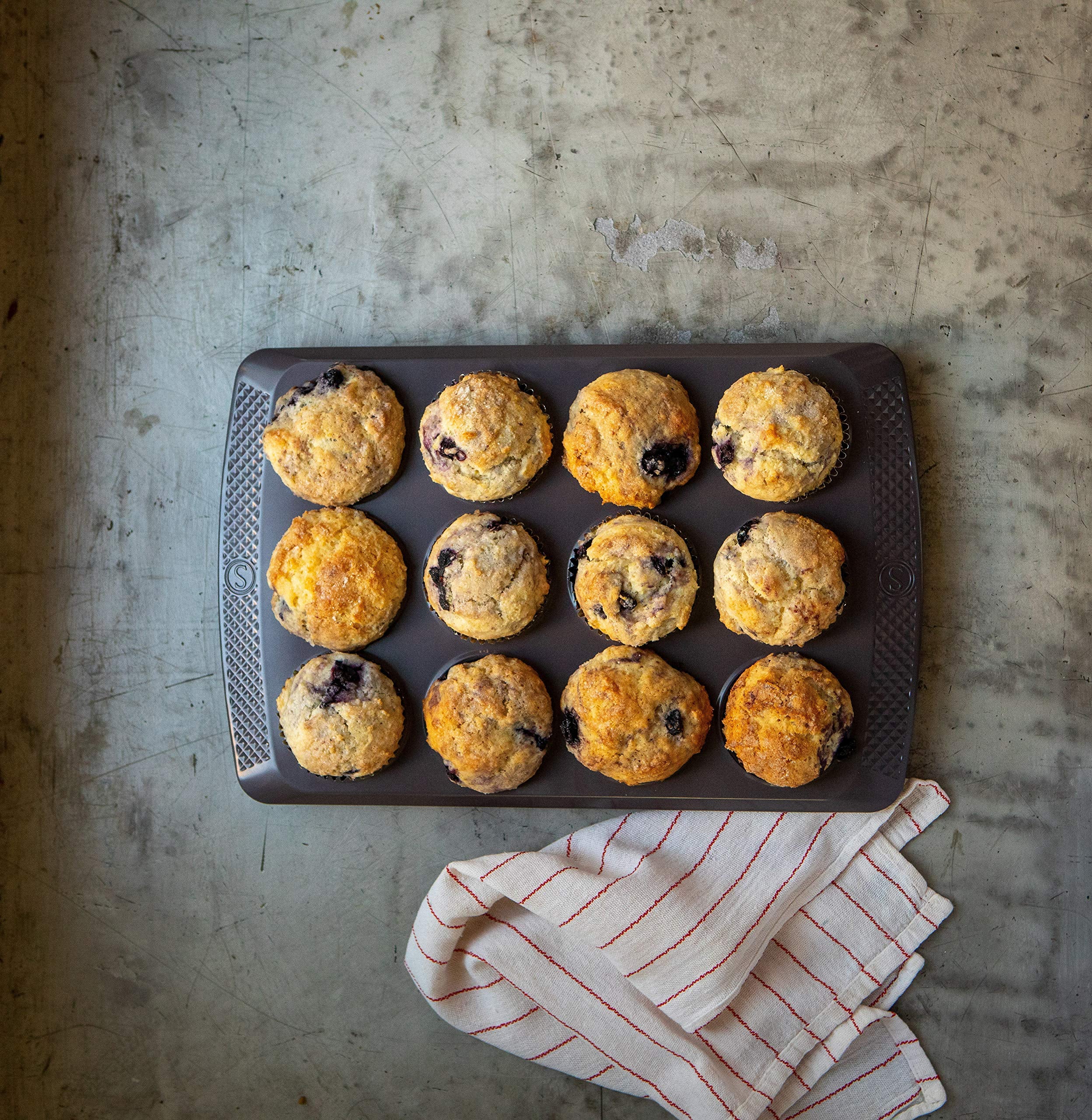 Muffins in a baking tray on a textured surface with a kitchen towel.