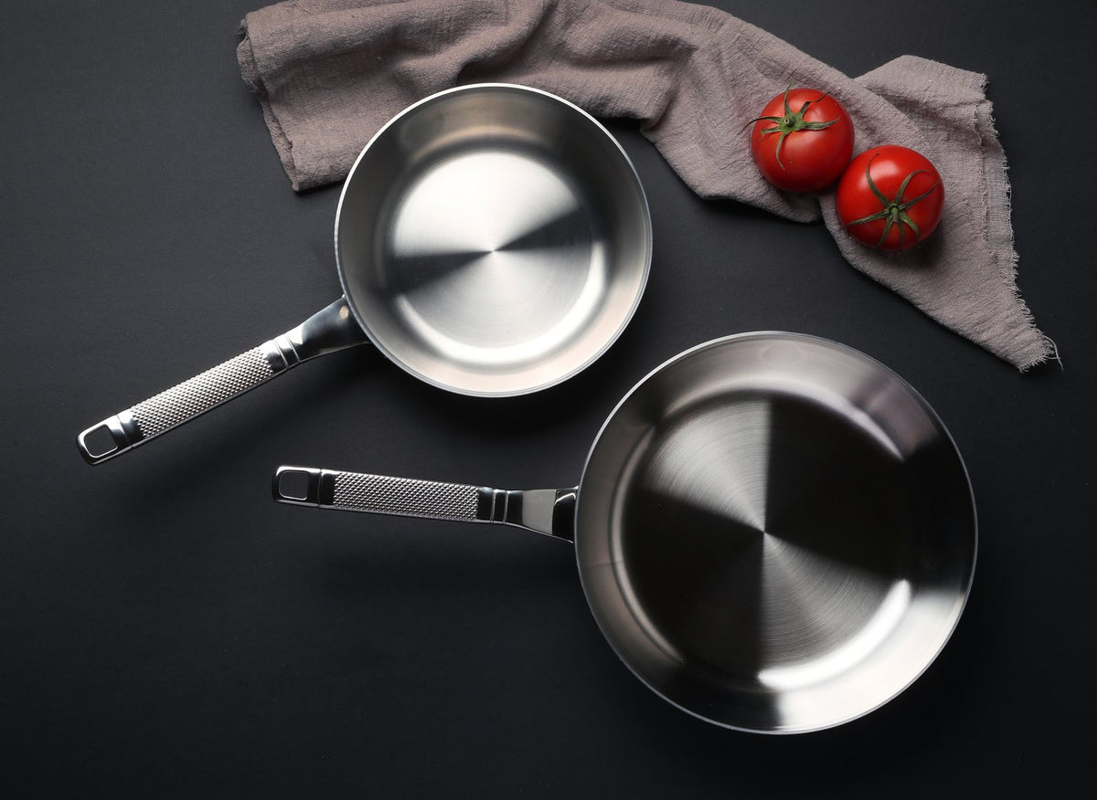 Two stainless steel frying pans on a dark surface with tomatoes and a cloth