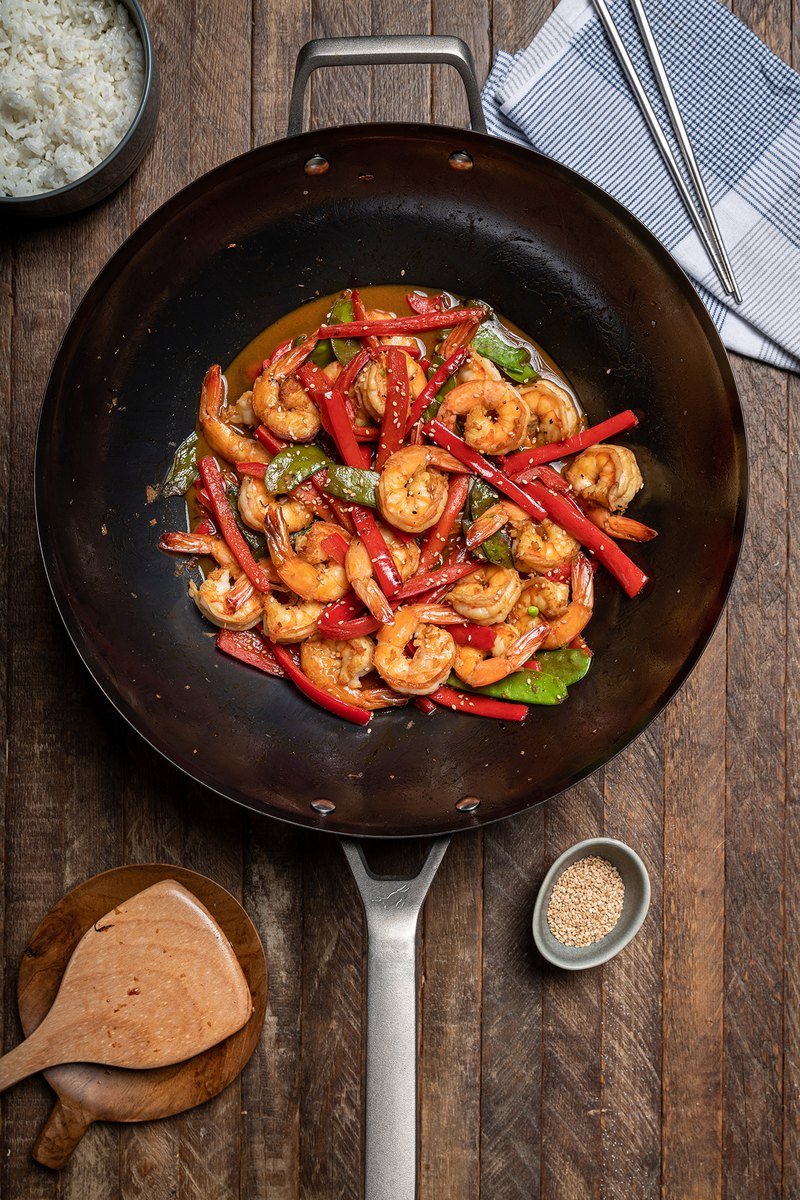A carbon steel wok filled with stir-fried vegetables and prawns on a wooden surface