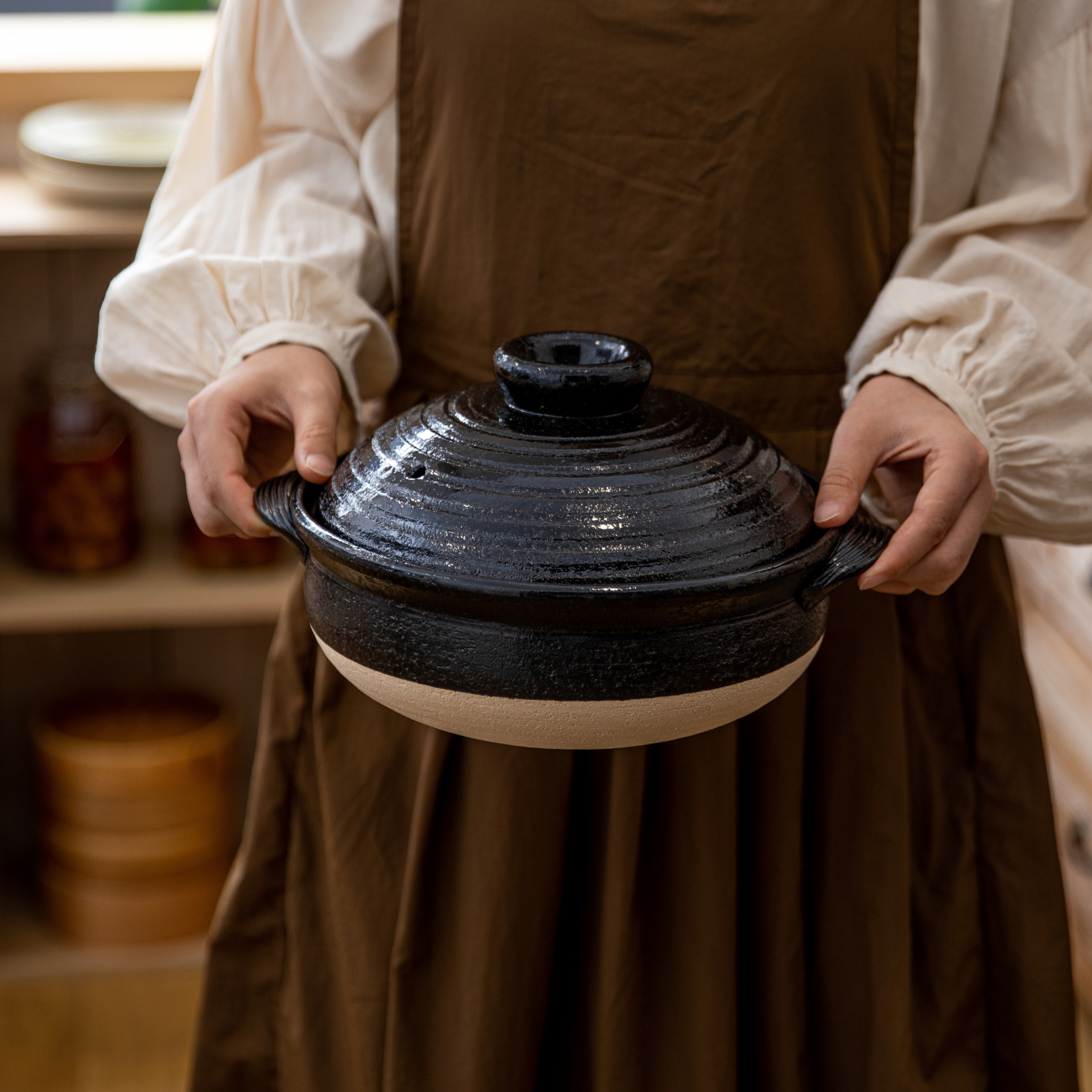 Person holding a black ceramic pot with a lid in a kitchen setting.
