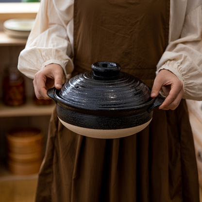 Person holding a black ceramic pot with a lid in a kitchen setting.